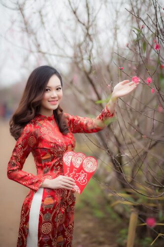 Young woman in traditional Vietnamese dress holding red envelopes in a blossoming garden.