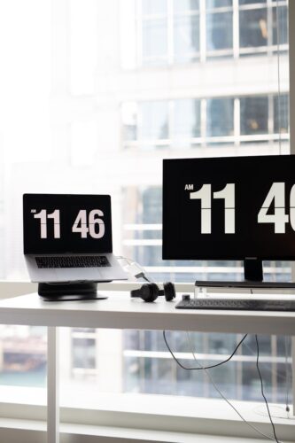 Minimalist modern office setup featuring a laptop and monitor displaying time against a corporate backdrop.