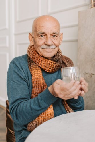 An elderly man with a mustache wearing a scarf smiles warmly while holding an empty glass inside a room.