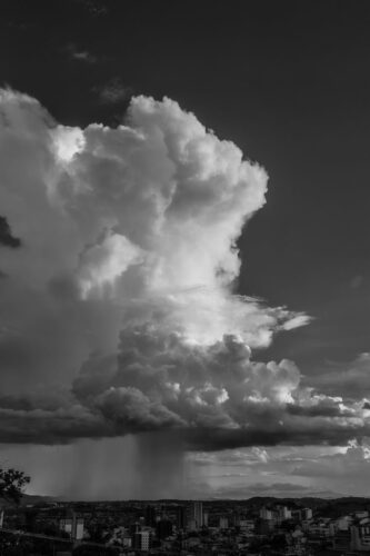 Black and white image of towering clouds with rain over a city, capturing dramatic weather change.
