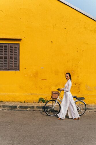 Asian woman in white dress walking a bicycle along a vibrant yellow wall on a sunny day.