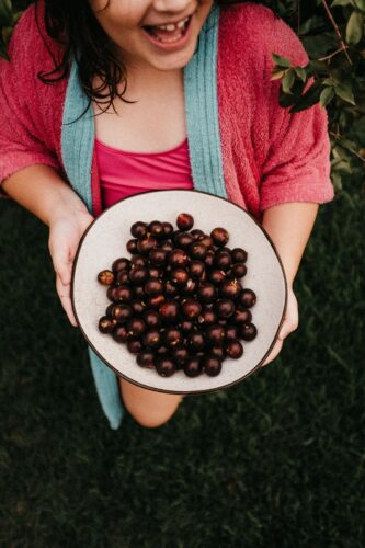 A joyful child holding a bowl of fresh jabuticaba fruits outdoors.