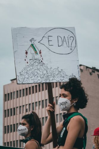 Women protesting for COVID-19 vaccine and human rights in Rio de Janeiro, holding placards and wearing face masks.