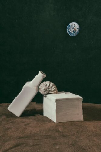 Surreal composition of a ceramic bottle and box on a textured surface with Earth in the background.