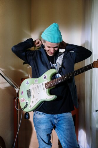 Musician wearing a beanie tunes electric guitar in a cozy home studio.