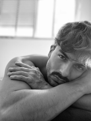 Artistic black and white portrait of a shirtless man resting indoors with a thoughtful gaze.